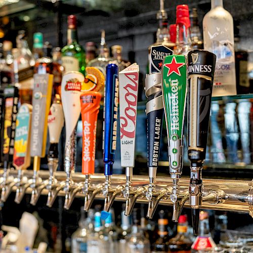 A row of beer taps with various brand labels in a bar setting, surrounded by bottles of liquor and glassware on shelves in the background.