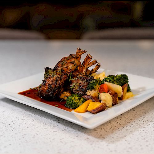 A gourmet dish featuring a rack of lamb served with roasted vegetables and garnishes, presented on a white square plate on a speckled countertop.