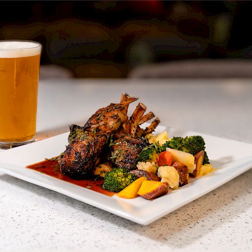 A plate of grilled lamb chops with vegetables and a glass of beer on a countertop.