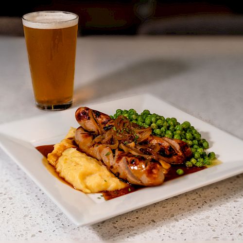 The image shows a plate with sausages, mashed potatoes, peas, and caramelized onions, served with a glass of beer on a white countertop.