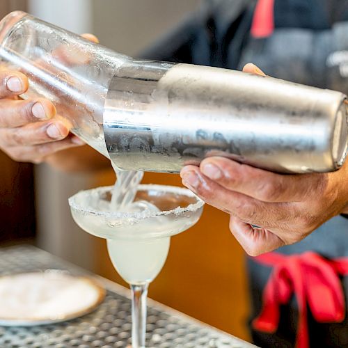 A bartender is pouring a cocktail from a shaker into a margarita glass with a salted rim, while standing at the bar.