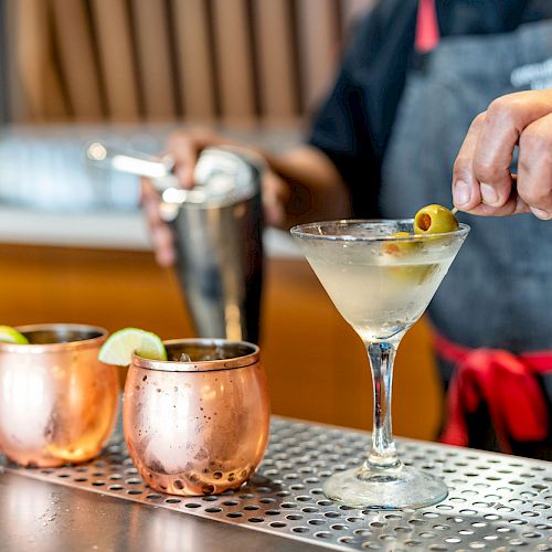 A bartender prepares a cocktail, garnishing a martini glass with an olive. Two Moscow mule mugs with lime slices sit nearby on the bar table.