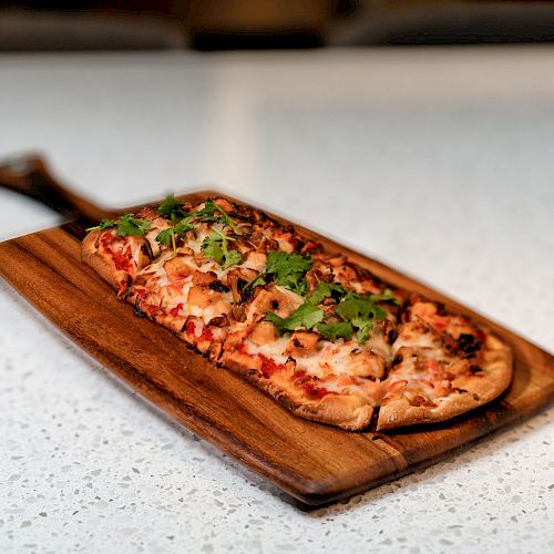 A flatbread pizza with toppings such as cheese, chicken, and cilantro is served on a wooden paddle, resting on a speckled countertop.