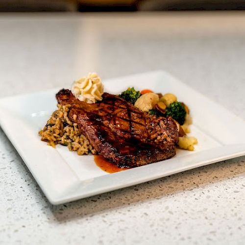 A dish of grilled steak on a bed of rice, served with a side of vegetables and garnished with a dollop of butter on a white square plate.