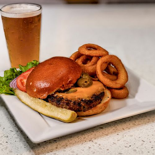 A burger with lettuce, tomato, onion, cheese, and jalapenos, served with a pickle and onion rings, accompanied by a glass of beer on a white plate.