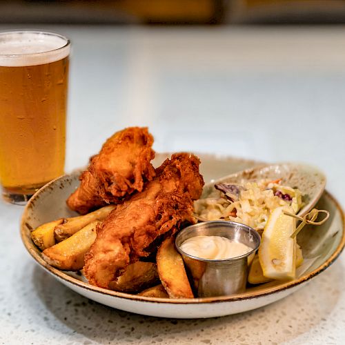 The image shows a plate with fried food, potato wedges, coleslaw, lemon, sauce, and a glass of beer.