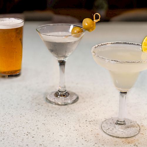 The image shows a pint of beer, a martini garnished with olives, and a margarita with a salted rim and a lime slice on a white surface.