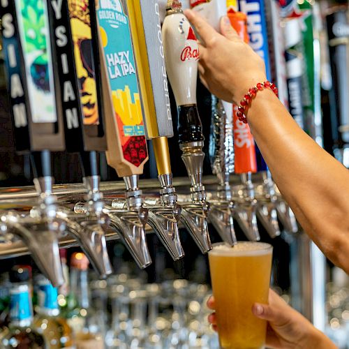 A person is pouring a beer from a tap handle into a glass at a bar, with various beer taps visible in the background.