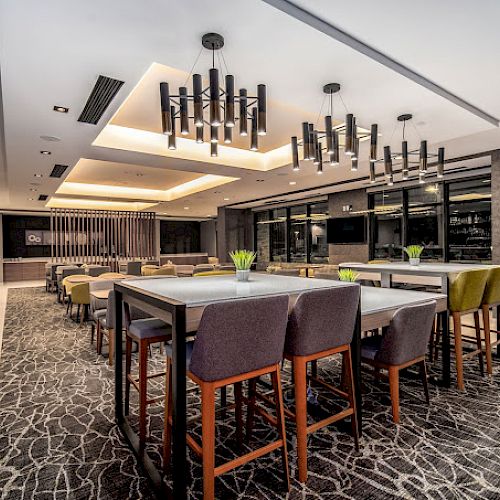 The image shows a modern dining area with high tables and cushioned chairs, stylish chandeliers, a patterned carpet, and a clean, contemporary design.