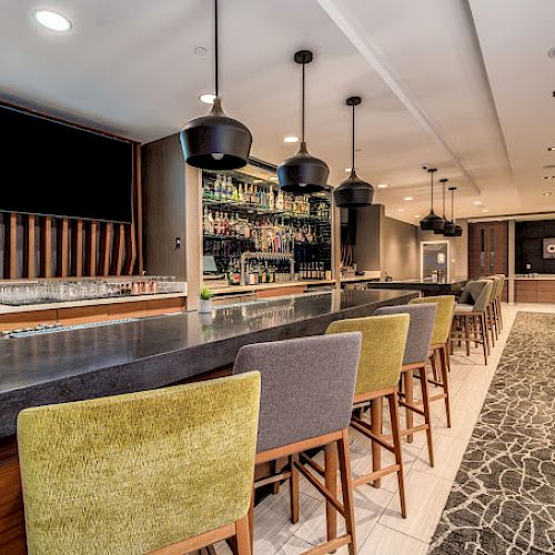 A modern bar with a sleek counter, bar stools, hanging lights, and a fully stocked back shelf next to a seating area with tables and chairs.