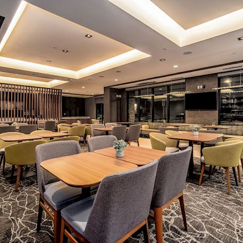 A modern dining area with wooden tables, grey and green upholstered chairs, patterned carpet, and ceiling lights, with large windows in the background.