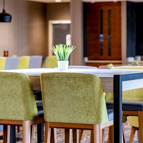 The image shows a modern dining area with green-upholstered chairs around a table, a small potted plant centerpiece, and pendant lights above.