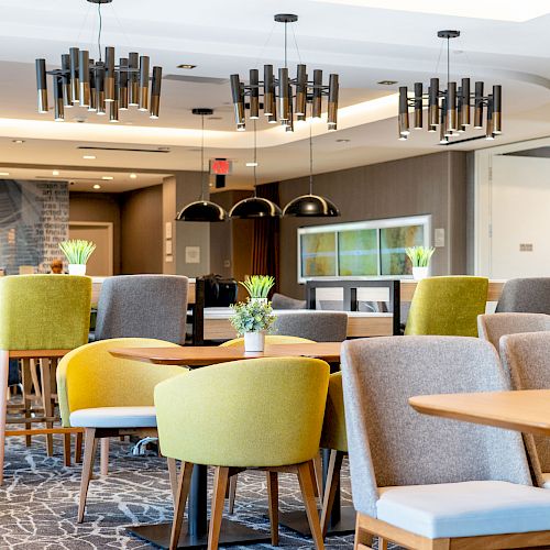 The image shows a modern dining area with green and gray chairs, wooden tables, and decorative hanging lights. Small potted plants are on the tables.