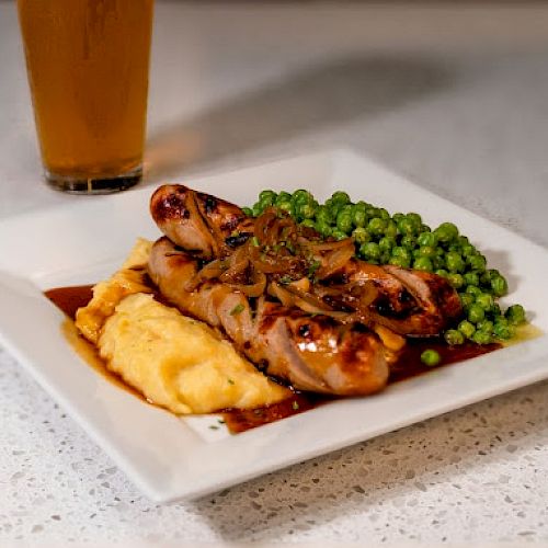 The image features a dish of sausages with mashed potatoes, peas, and onions on a white plate, along with a glass of beer in the background.