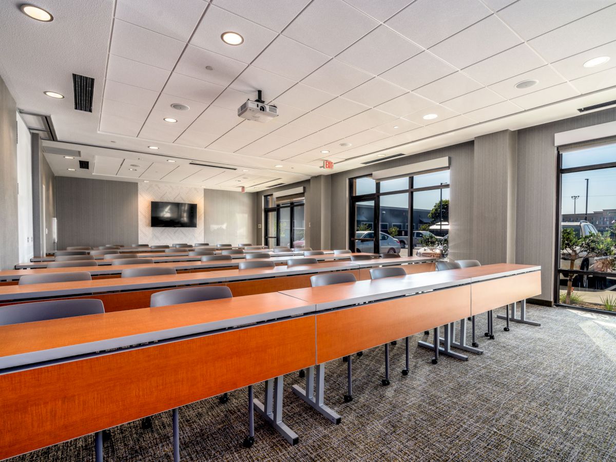 A modern conference room with long tables, gray chairs, a projector, and large windows allowing natural light to fill the space.