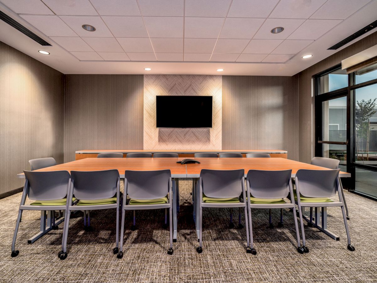 A modern conference room featuring a long table, several chairs, a wall-mounted TV screen, and large windows with a view of the outdoors.