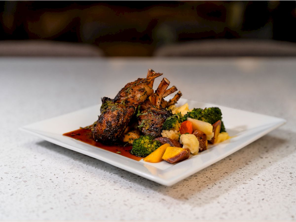 A plate of grilled lamb chops with a sauce, accompanied by broccoli, potatoes, and yellow squash on a white dish.