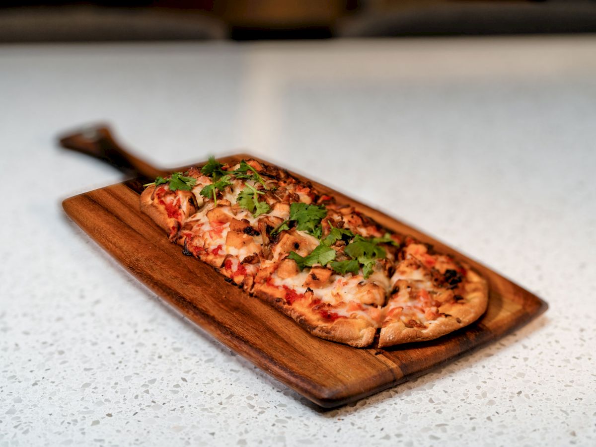 A flatbread pizza topped with cheese, vegetables, and herbs, is served on a wooden board placed on a white speckled countertop.