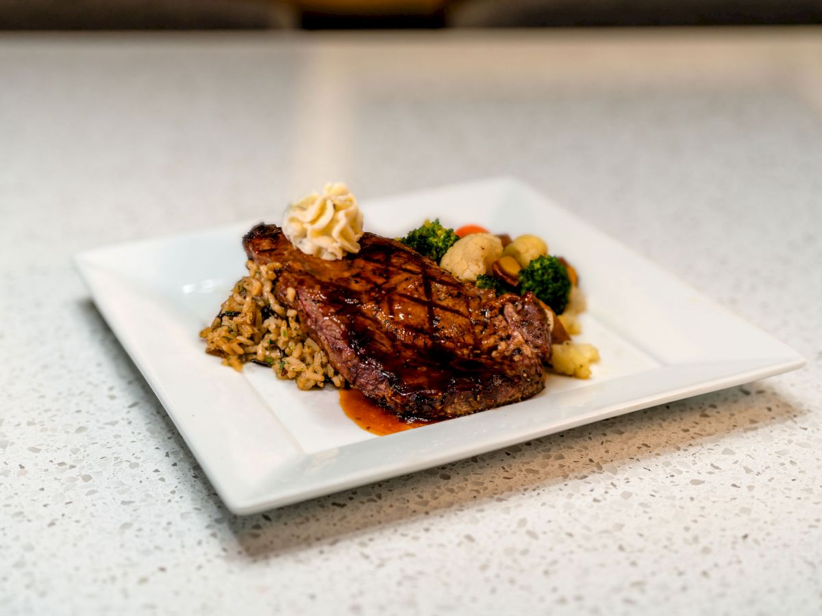 A grilled steak is served with rice, vegetables, and a dollop of butter on a square white plate atop a white speckled counter.