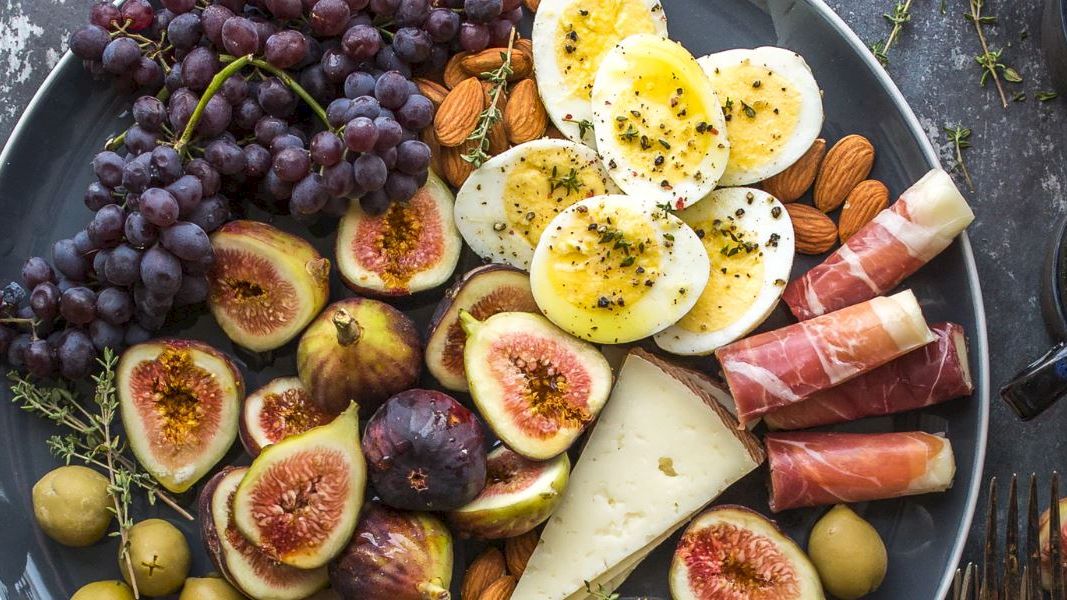A platter featuring figs, grapes, boiled eggs, prosciutto, cheese, almonds, olives with two cups of coffee and a fork on the side ending the sentence.