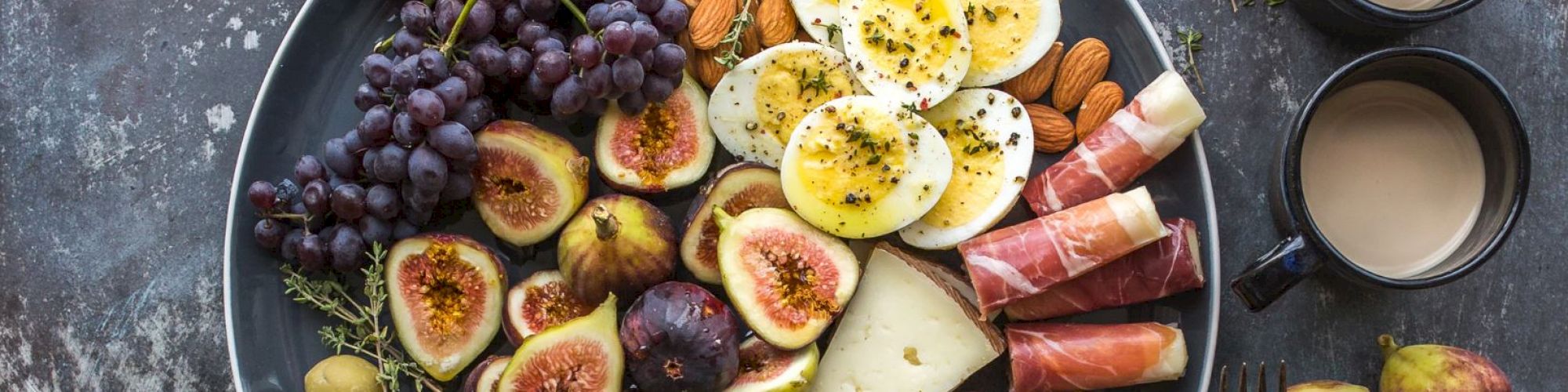 A platter featuring figs, grapes, boiled eggs, prosciutto, cheese, almonds, olives with two cups of coffee and a fork on the side ending the sentence.