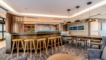 The image shows a modern lounge area with bar stools, a counter, a table with chairs, and contemporary lighting and decor.