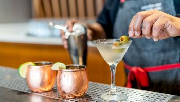 A bartender is garnishing a martini with an olive, with two copper mugs containing drinks with lime wedges on the side.