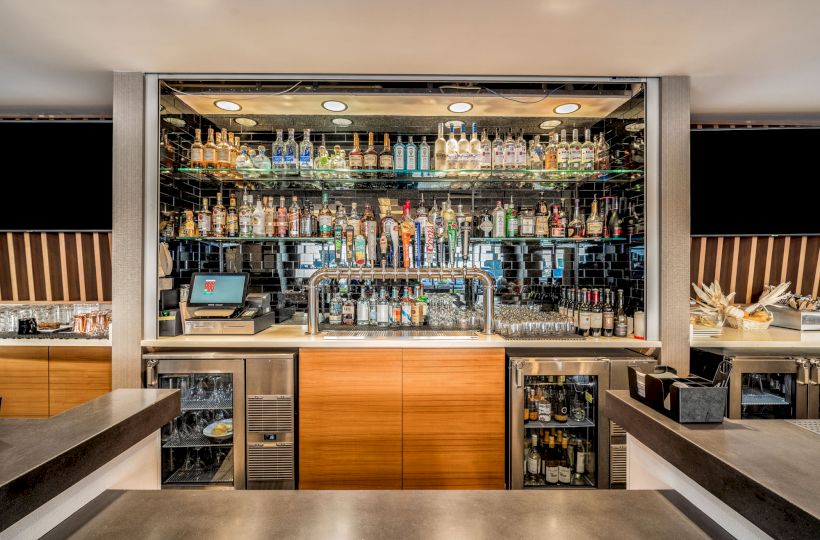 The image shows a well-organized bar with a variety of liquor bottles, a cash register, taps for draft beer, and bar tools on the counter.