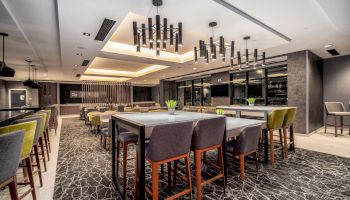 The image depicts a modern dining area with high tables and chairs, contemporary lighting fixtures, and decorative elements like potted plants.