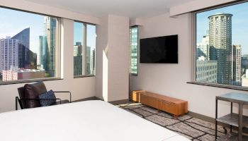 The image is a modern hotel room with large windows showing a cityscape, a TV on the wall, and minimalistic furniture.