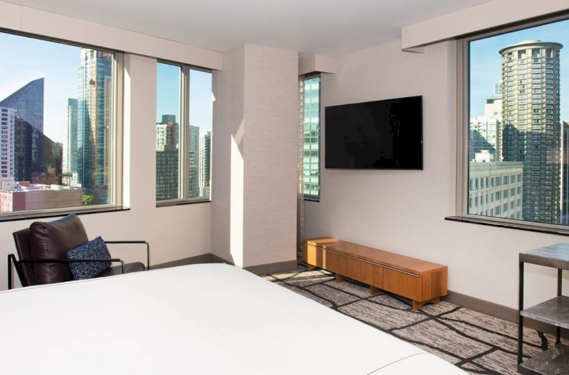 The image is a modern hotel room with large windows showing a cityscape, a TV on the wall, and minimalistic furniture.