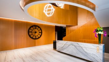 The image depicts a modern lobby with wooden accents, a large wall clock, a marble reception desk, and decorative lighting and flowers.