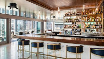 The image shows a modern bar with a wooden counter, blue and gold bar stools, and shelves filled with various bottles of alcohol, with city views outside.