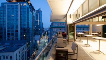 The image shows a modern balcony with seating, tables, and cityscape views. High-rise buildings and a street are visible in the background.