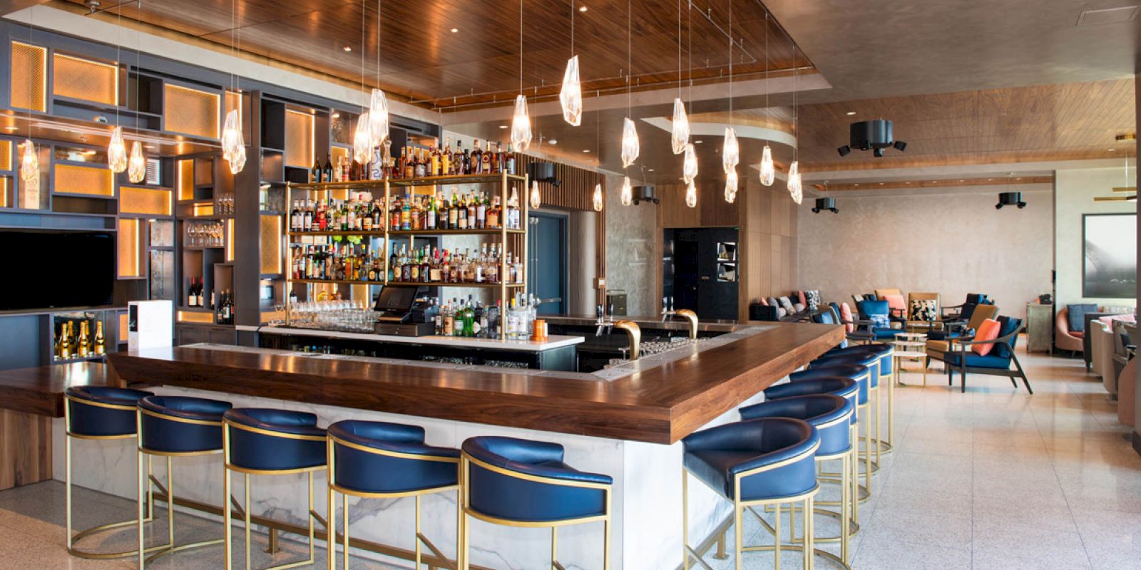 The image shows a modern bar with a wooden countertop, blue stools, a well-stocked back bar, hanging lights, and a lounge area in the background.