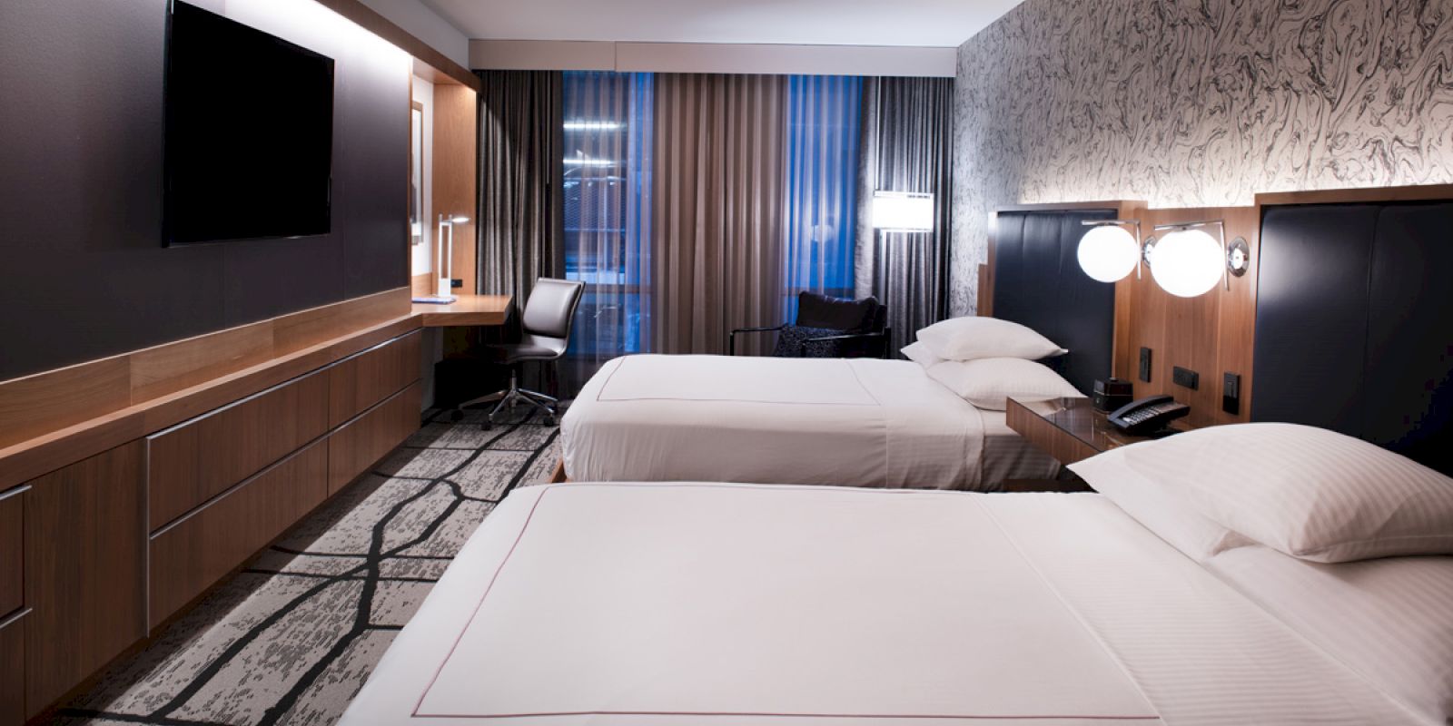 A modern hotel room with two beds, a wall-mounted TV, a desk with a chair, and contemporary lighting and decor.
