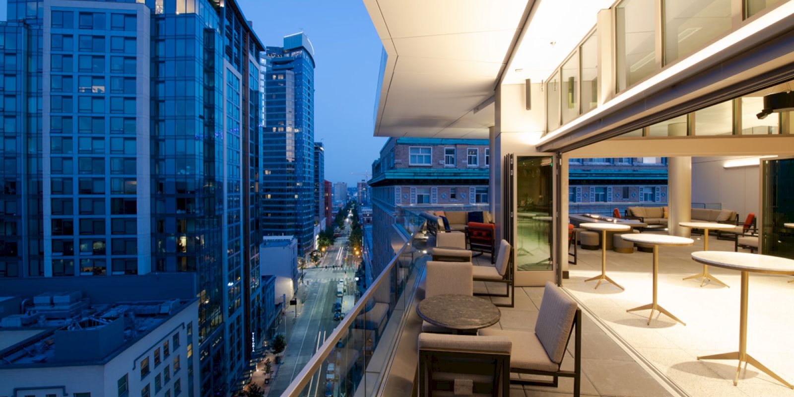 The image shows a modern rooftop terrace with tables and chairs. The terrace overlooks a city street with tall buildings at dusk.