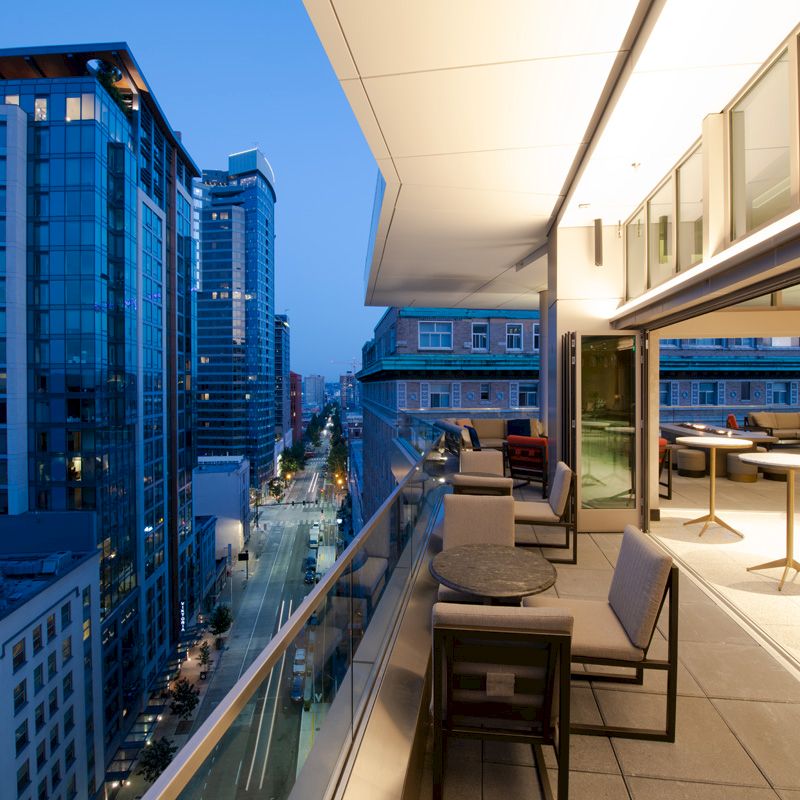 The image shows a modern cityscape at dusk with a view from a high-rise balcony furnished with chairs and tables, overlooking illuminated buildings.