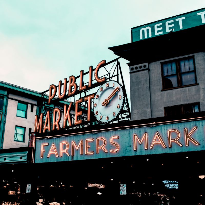 The image shows a neon sign for a Public Market and Farmers Market on an urban street, with buildings in the background displaying additional signage.