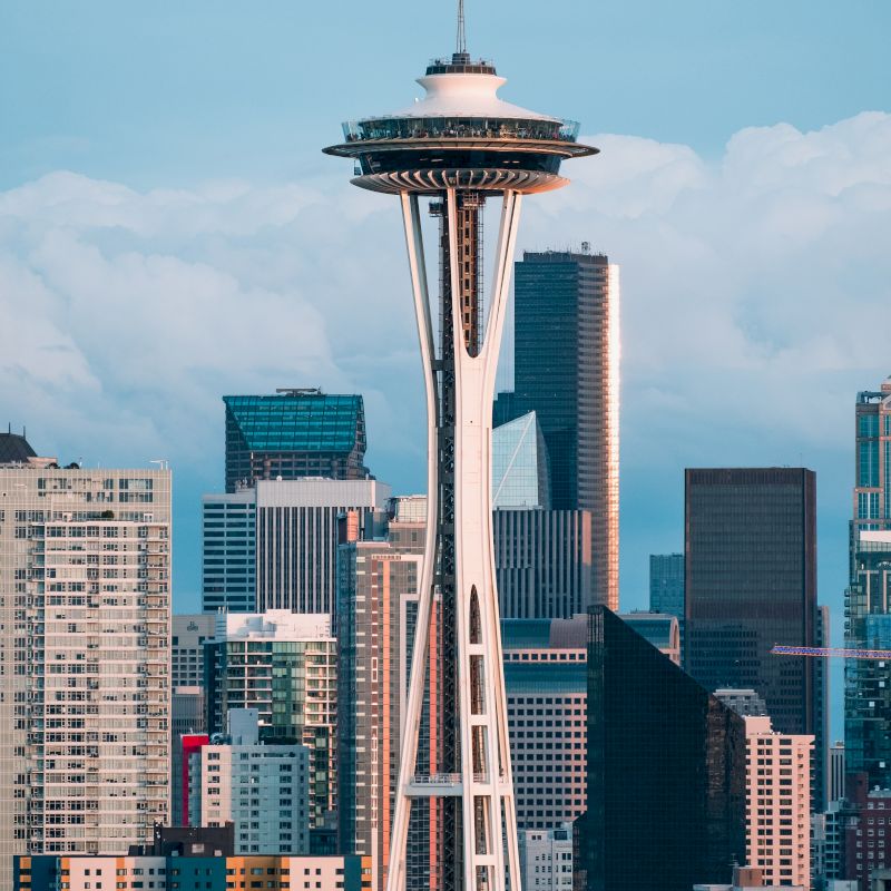 The image features the iconic Space Needle in Seattle, with the downtown city's skyline and various skyscrapers in the background, under a partly cloudy sky.