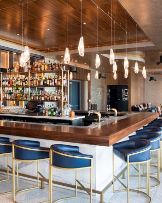 A modern bar area with blue bar stools, hanging lights, and a well-stocked bar. The space features wooden accents and a seating area in the background.