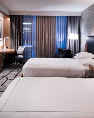 The image shows a modern hotel room with two neatly made double beds, a large TV, a desk with a chair, and a well-lit window with curtains.