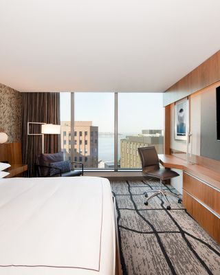 This image shows a modern hotel room with a large bed, a flat-screen TV, a desk, and a view of city buildings through large windows.