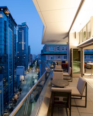The image shows a modern, upscale outdoor balcony with tables and chairs, overlooking a cityscape with tall buildings and a street below.