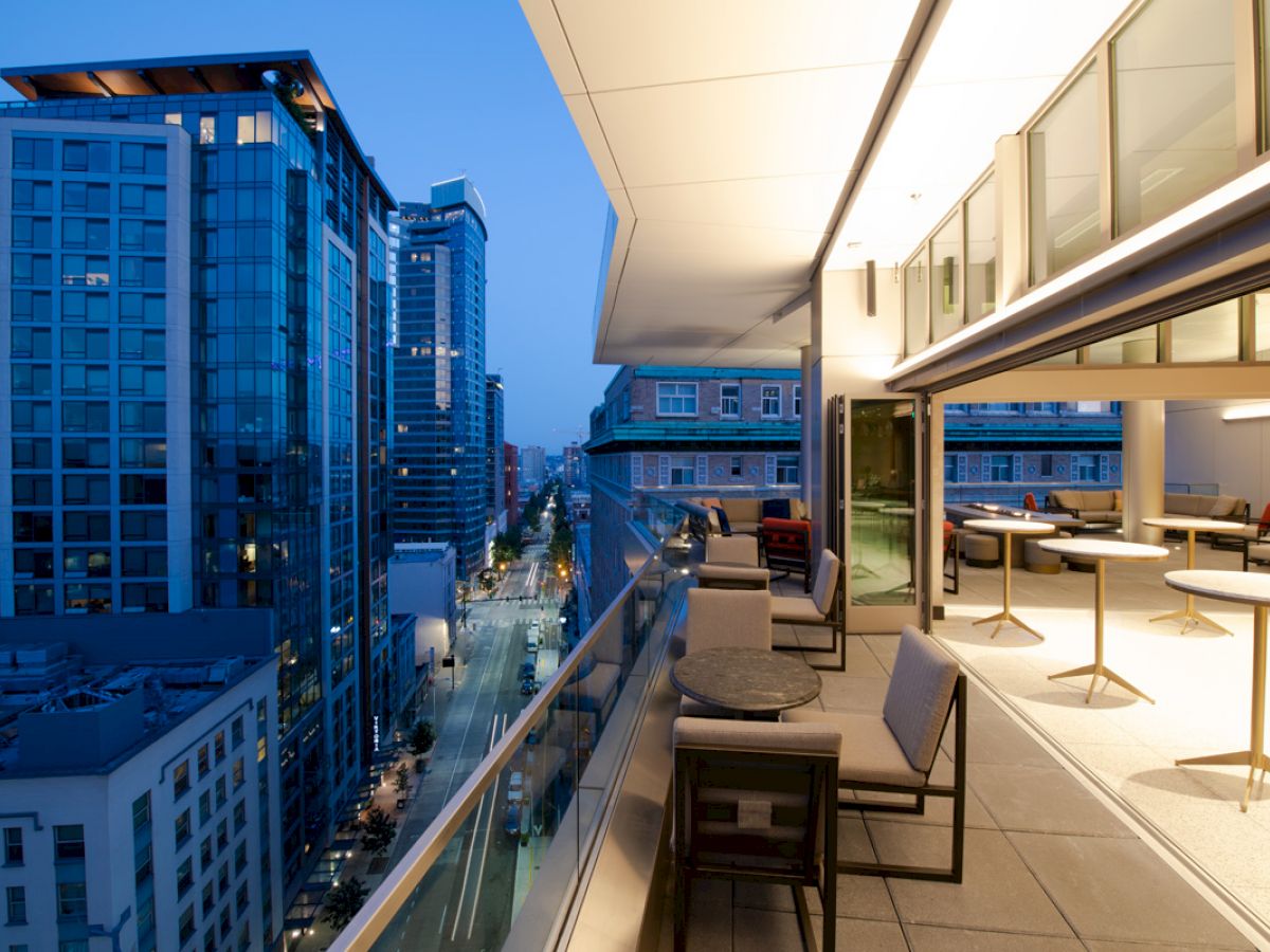 This image shows a cityscape with tall buildings and a modern high-rise balcony at dusk. The balcony has tables, chairs, and lighting.