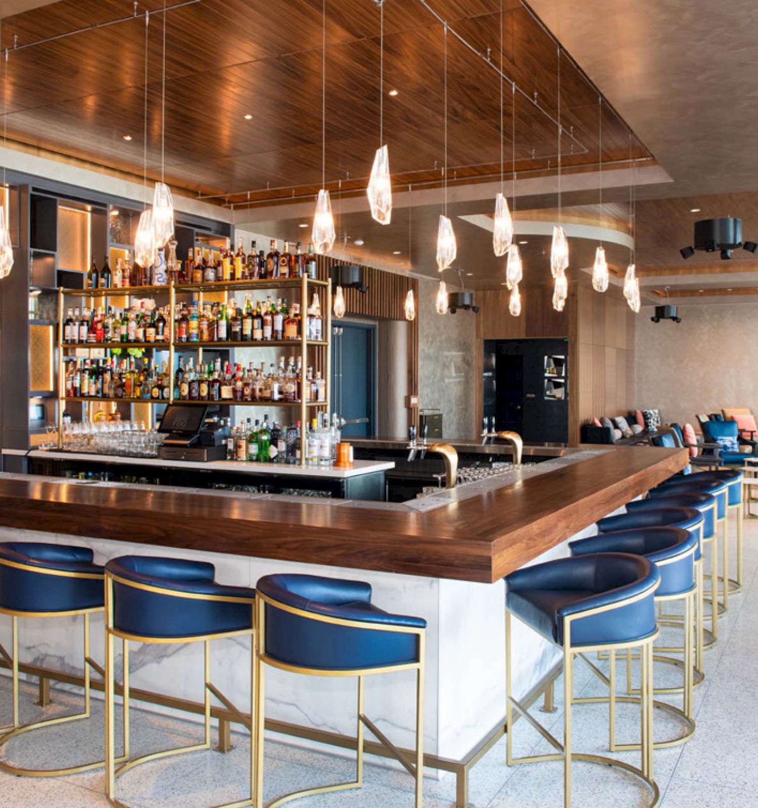 The image shows a modern bar with a wooden counter, blue cushioned stools, hanging lights, and a well-stocked bar area in a spacious, stylish setting.