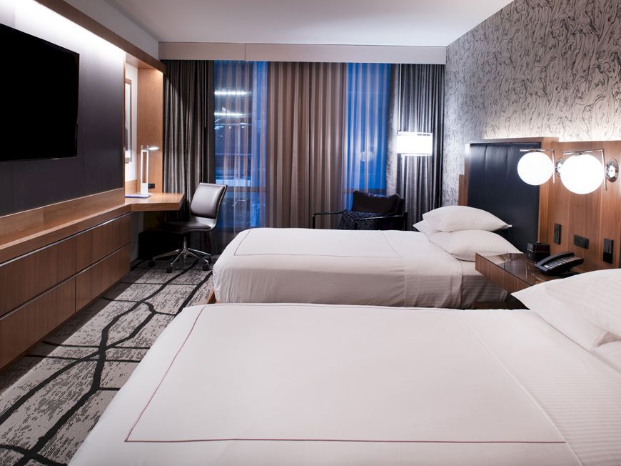 A hotel room with two beds, modern decor, a wall-mounted TV, a work desk, lamps, curtains, and patterned carpeting.