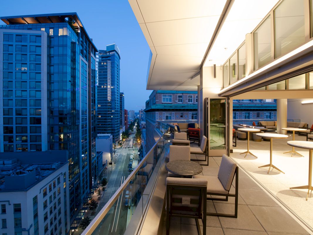 A modern cityscape with tall buildings and a view from a stylish balcony featuring tables, chairs, and an indoor lounge area at dusk.