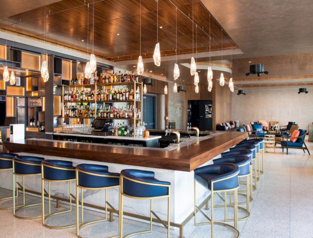 A modern bar features a well-stocked shelf, sleek barstools, hanging lights, and a cozy seating area with couches and tables in the background.