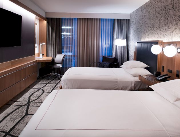 A modern hotel room features two beds, a desk with a chair, wall-mounted TV, nightstands with lamps, and large windows with curtains.